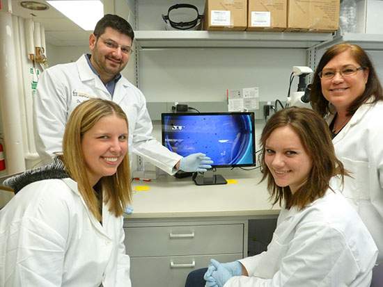 Four people in lab coats and gloves standing in front of a computer screen smiling