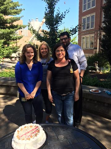 Four people standing outside with a cake on a table in front of them